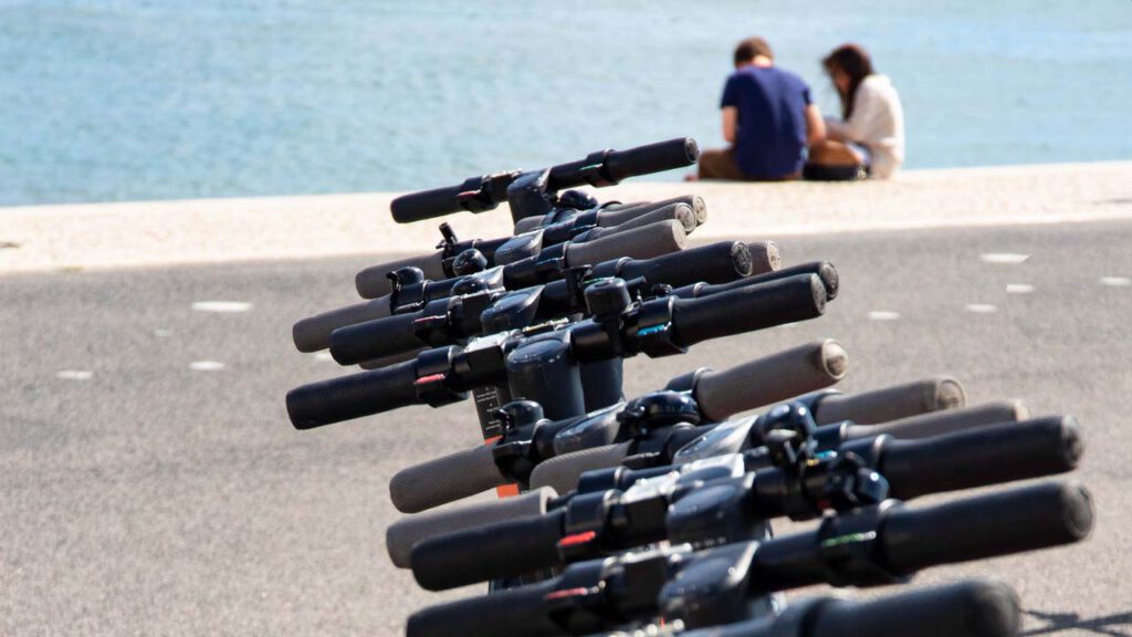E.Scooter stehen am Wasser, ein paar im Hintergrund unterhält sich
